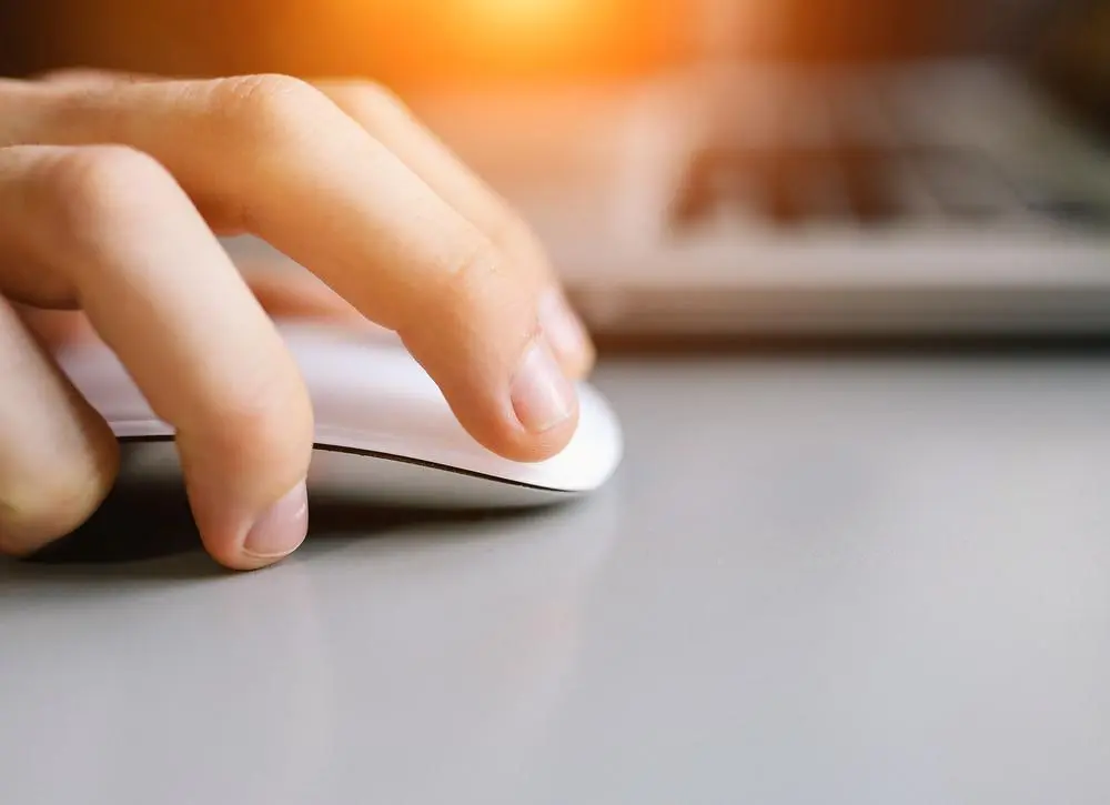 A close-up of a hand using a white computer mouse on a gray surface, with a blurred laptop keyboard in the background and warm light shining from above.