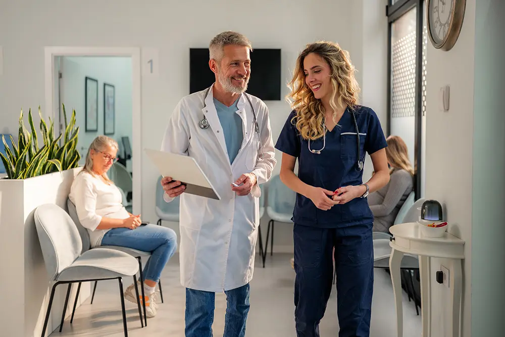 A male doctor and a female nurse walk and talk together in a bright medical clinic, while two patients sit in the waiting area in the background.