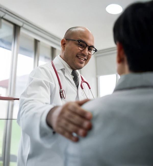 A smiling doctor wearing glasses and a white coat with a stethoscope gently places a hand on a patient’s shoulder in a bright medical office.